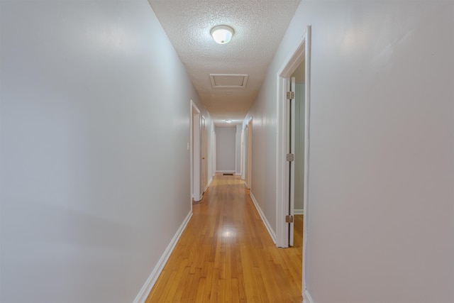 hall featuring attic access, light wood-type flooring, a textured ceiling, and baseboards