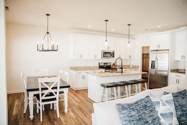 kitchen with appliances with stainless steel finishes, light wood-type flooring, pendant lighting, white cabinetry, and an island with sink