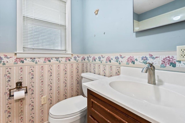 half bath featuring toilet, wallpapered walls, vanity, and wainscoting