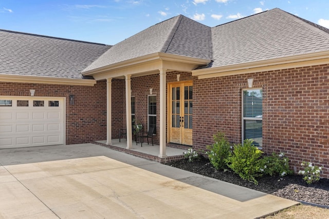 property entrance with driveway, french doors, brick siding, an attached garage, and a shingled roof