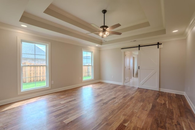 empty room featuring wood-type flooring, ceiling fan, a tray ceiling, crown molding, and a barn door