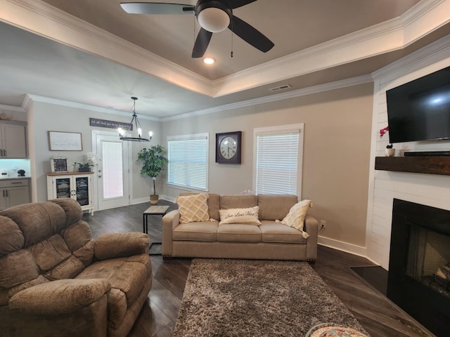 living room featuring ornamental molding, ceiling fan with notable chandelier, a large fireplace, a tray ceiling, and dark wood-type flooring