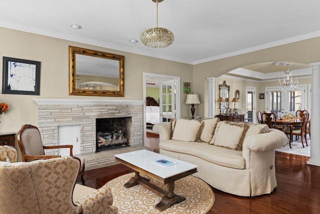 living room with a stone fireplace, crown molding, a notable chandelier, wood-type flooring, and decorative columns