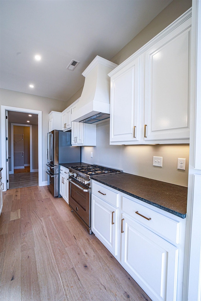 kitchen with custom exhaust hood, dark stone counters, white cabinets, high end stainless steel range oven, and light wood-type flooring