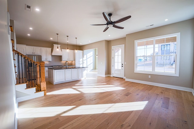 kitchen featuring custom range hood, a center island with sink, white cabinets, light hardwood / wood-style floors, and hanging light fixtures