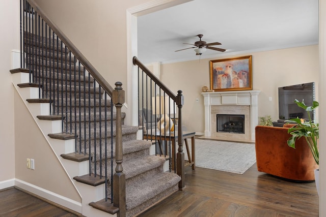stairs featuring wood finished floors, a ceiling fan, a premium fireplace, baseboards, and ornamental molding