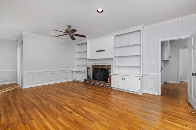 unfurnished living room featuring built in features, ceiling fan, crown molding, a brick fireplace, and light wood-type flooring