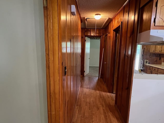 corridor with light wood finished floors, wood walls, attic access, and a textured ceiling