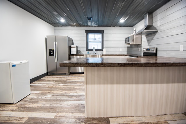 kitchen with sink, kitchen peninsula, stainless steel appliances, wall chimney range hood, and light hardwood / wood-style flooring