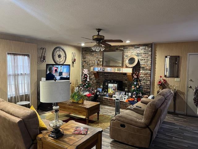 living room with ceiling fan, wood walls, a textured ceiling, and a brick fireplace