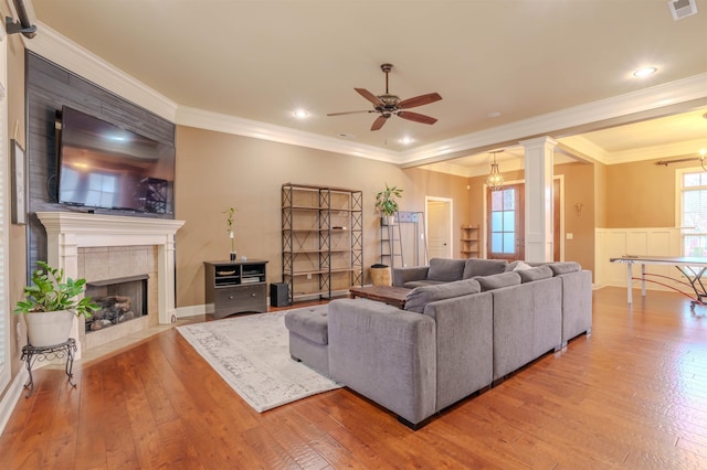 living room with crown molding, wood-type flooring, a tiled fireplace, and ornate columns