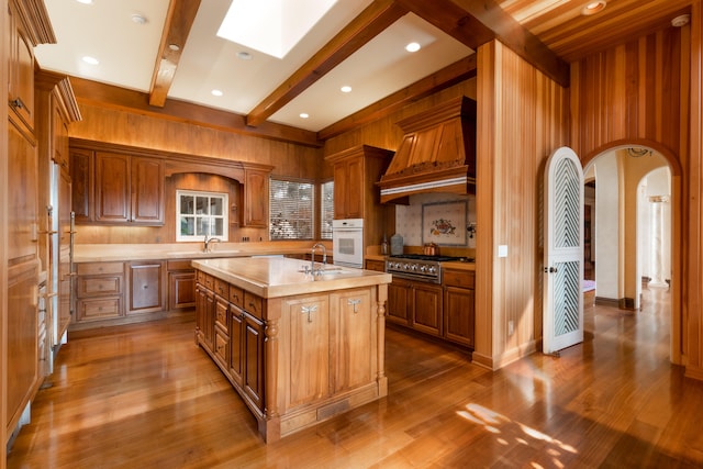 kitchen featuring an island with sink, premium range hood, stovetop, oven, and dark hardwood / wood-style flooring