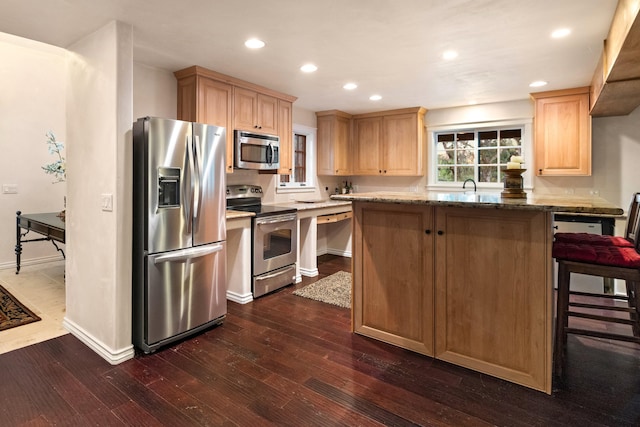 kitchen with light brown cabinets, appliances with stainless steel finishes, a kitchen bar, dark wood-type flooring, and dark stone counters