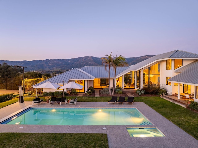 pool at dusk with an in ground hot tub, a mountain view, a yard, and a patio area