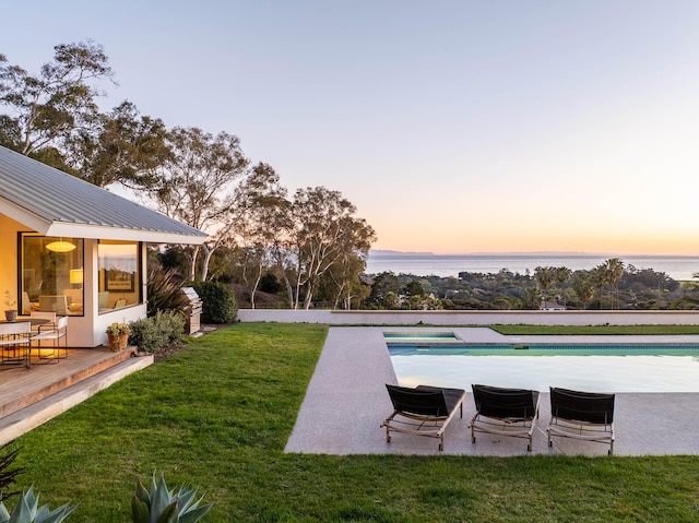 pool at dusk featuring a deck with water view and a lawn