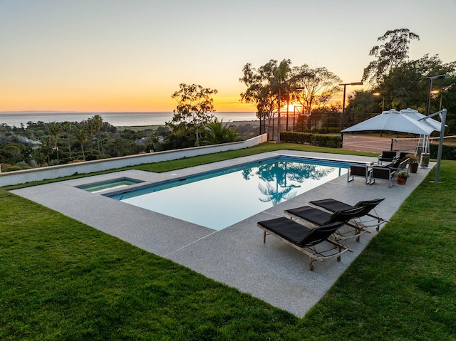 pool at dusk with an in ground hot tub, a water view, a patio area, and a lawn