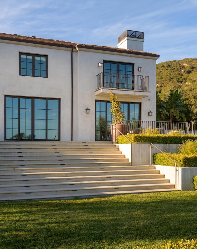 rear view of property with a balcony, a yard, stairway, stucco siding, and a chimney