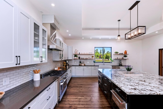 kitchen with wall chimney range hood, dark stone countertops, decorative light fixtures, range with two ovens, and white cabinets