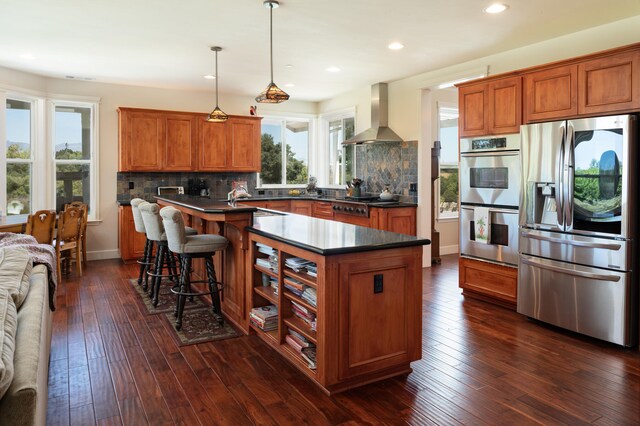 kitchen with appliances with stainless steel finishes, wall chimney range hood, pendant lighting, decorative backsplash, and dark hardwood / wood-style flooring