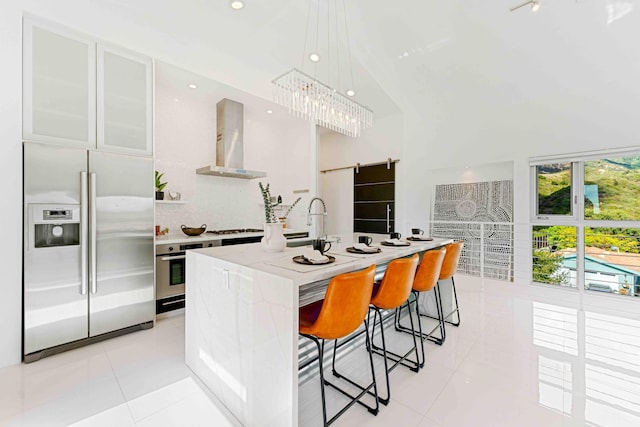 kitchen featuring a center island with sink, stainless steel appliances, white cabinetry, a barn door, and wall chimney exhaust hood