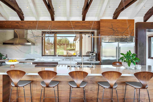 kitchen featuring beamed ceiling, sink, wall chimney range hood, a kitchen breakfast bar, and stainless steel built in fridge