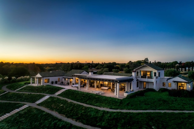 back house at dusk featuring a balcony, a lawn, and a patio