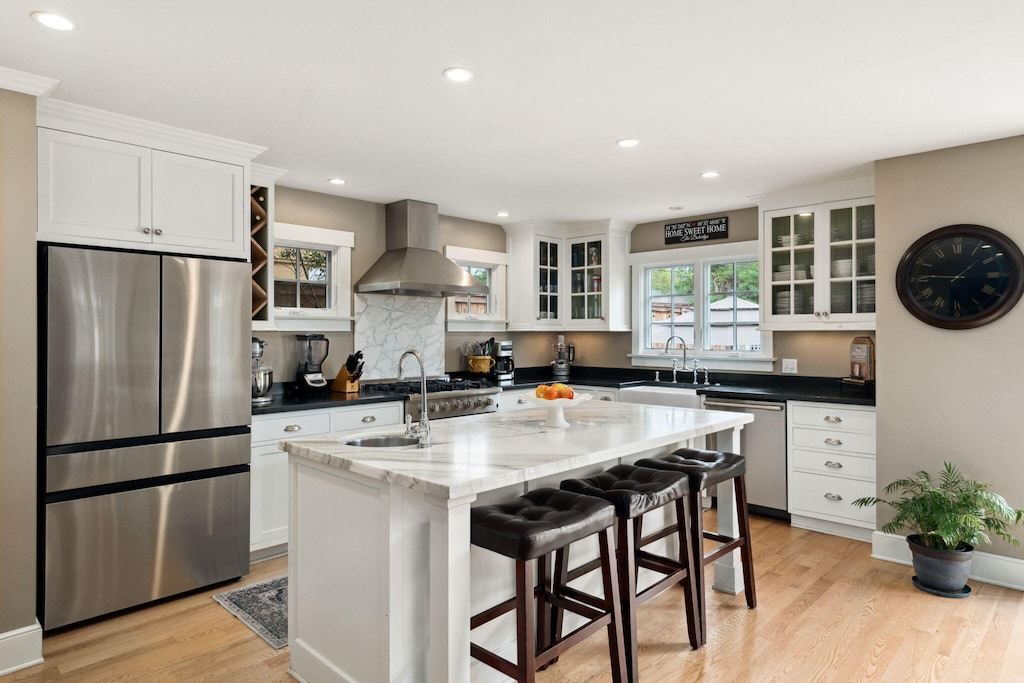 kitchen featuring wall chimney range hood, light hardwood / wood-style flooring, an island with sink, appliances with stainless steel finishes, and white cabinetry