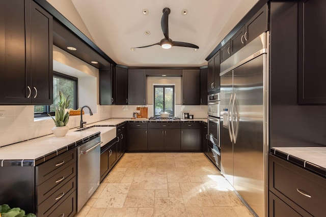 kitchen with tile countertops, vaulted ceiling, sink, backsplash, and appliances with stainless steel finishes