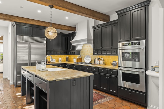 kitchen with hanging light fixtures, premium range hood, backsplash, an island with sink, and appliances with stainless steel finishes