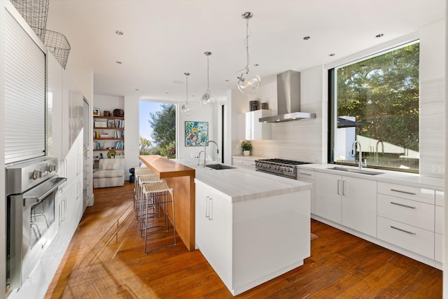 kitchen with oven, sink, wall chimney exhaust hood, an island with sink, and white cabinetry