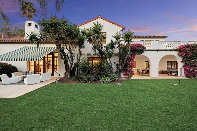 back house at dusk with a yard, a balcony, a patio, and an outdoor hangout area