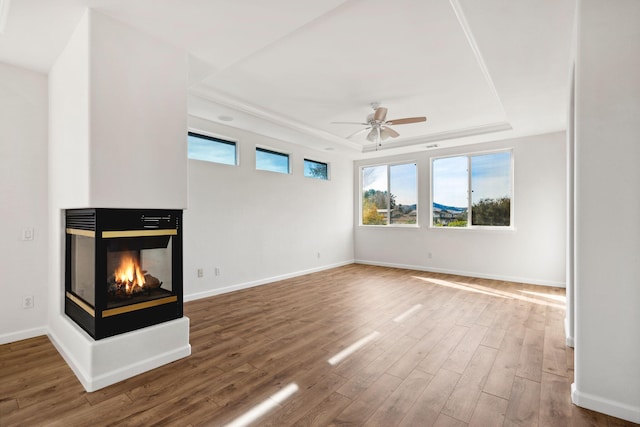 unfurnished living room featuring ceiling fan, wood finished floors, a multi sided fireplace, baseboards, and a raised ceiling