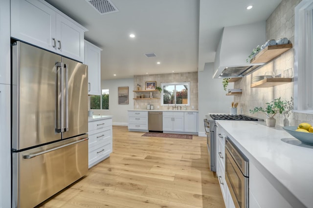 kitchen with light wood-type flooring, backsplash, premium appliances, ventilation hood, and white cabinets