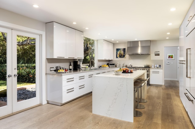 kitchen featuring a center island, wall chimney range hood, a kitchen breakfast bar, white cabinets, and light wood-type flooring