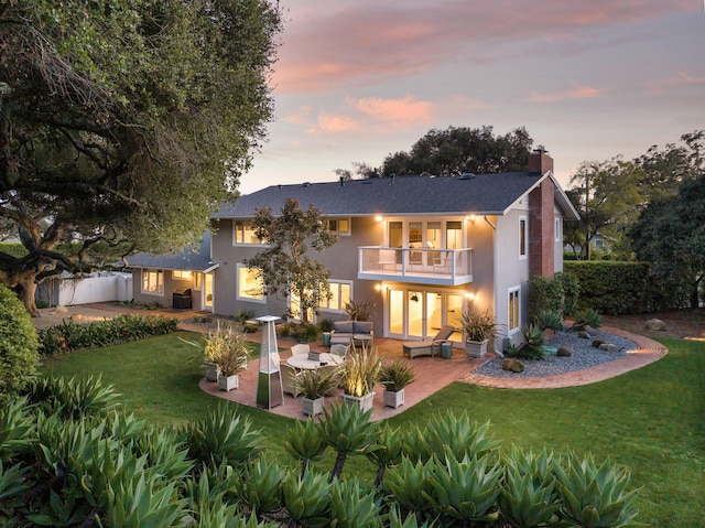 back house at dusk featuring outdoor lounge area, a balcony, a patio, and a lawn