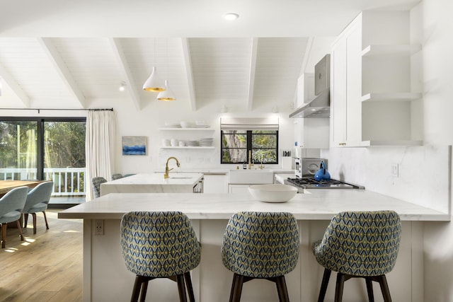 kitchen featuring a breakfast bar, white cabinetry, light stone counters, beamed ceiling, and wall chimney range hood