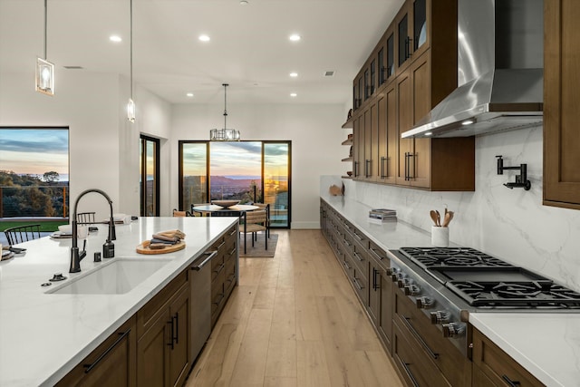 kitchen featuring wall chimney exhaust hood, appliances with stainless steel finishes, glass insert cabinets, hanging light fixtures, and a sink