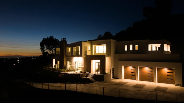 back of house at dusk with a garage, driveway, fence, and stucco siding