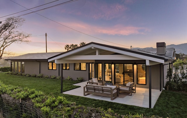 back of property at dusk featuring a chimney, outdoor lounge area, a lawn, a patio, and stucco siding