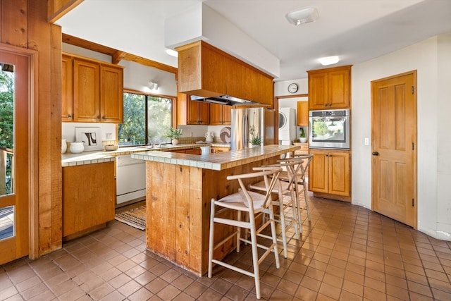 kitchen featuring tile countertops, tile patterned floors, stacked washing maching and dryer, an island with sink, and stainless steel appliances