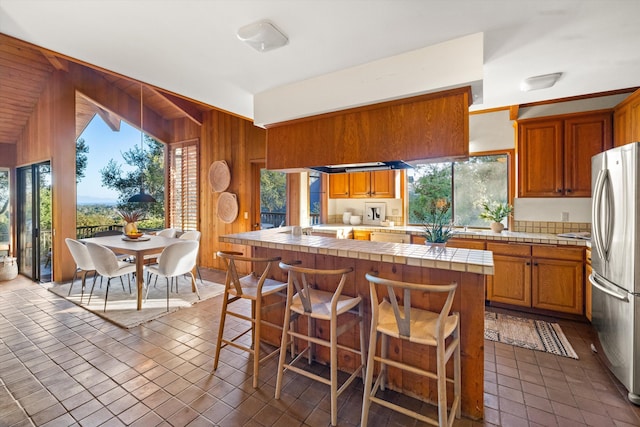 kitchen with wooden walls, tile counters, a kitchen island, plenty of natural light, and stainless steel refrigerator