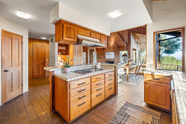 kitchen featuring kitchen peninsula, tile countertops, wood walls, stainless steel gas stovetop, and range hood