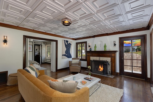 living area featuring dark wood-style flooring, baseboards, a tiled fireplace, an ornate ceiling, and crown molding