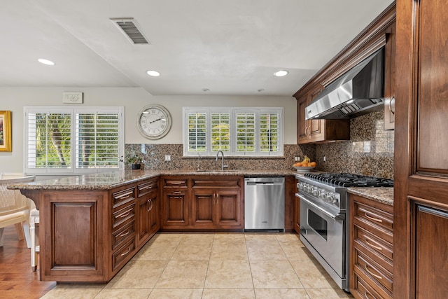 kitchen with wall chimney exhaust hood, sink, dark stone countertops, kitchen peninsula, and stainless steel appliances