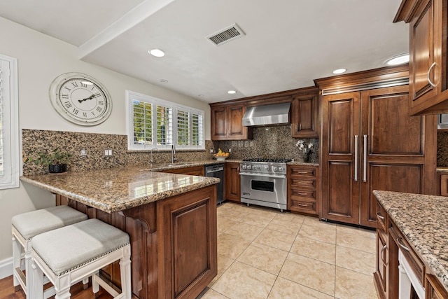 kitchen featuring wall chimney exhaust hood, a kitchen breakfast bar, high end stainless steel range oven, light stone countertops, and decorative backsplash