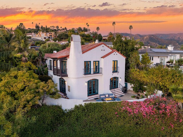 back of house at dusk with stucco siding, french doors, a chimney, a balcony, and a patio