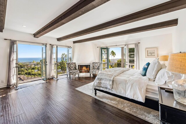 bedroom with wood finished floors, baseboards, beam ceiling, a warm lit fireplace, and access to exterior