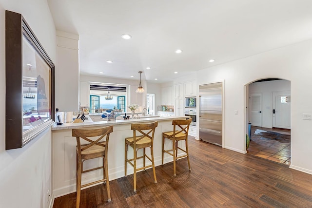 kitchen with a peninsula, dark wood-style flooring, arched walkways, white cabinets, and built in appliances