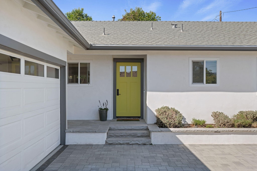 entrance to property featuring a garage, roof with shingles, and stucco siding