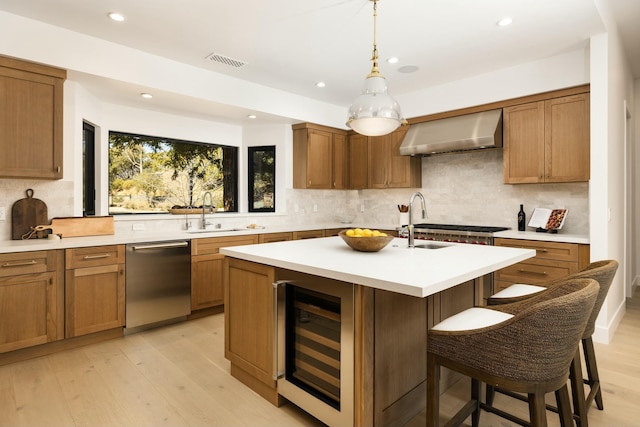 kitchen with wall chimney exhaust hood, sink, appliances with stainless steel finishes, an island with sink, and beverage cooler